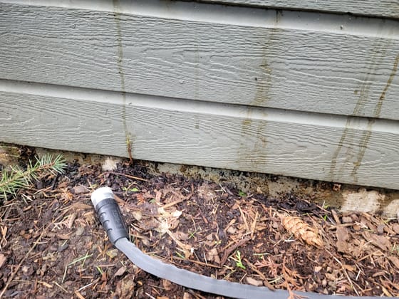 Water staining running down gray siding to a darkened, decaying bottom edge where it meets the foundation, mulch and a hose below